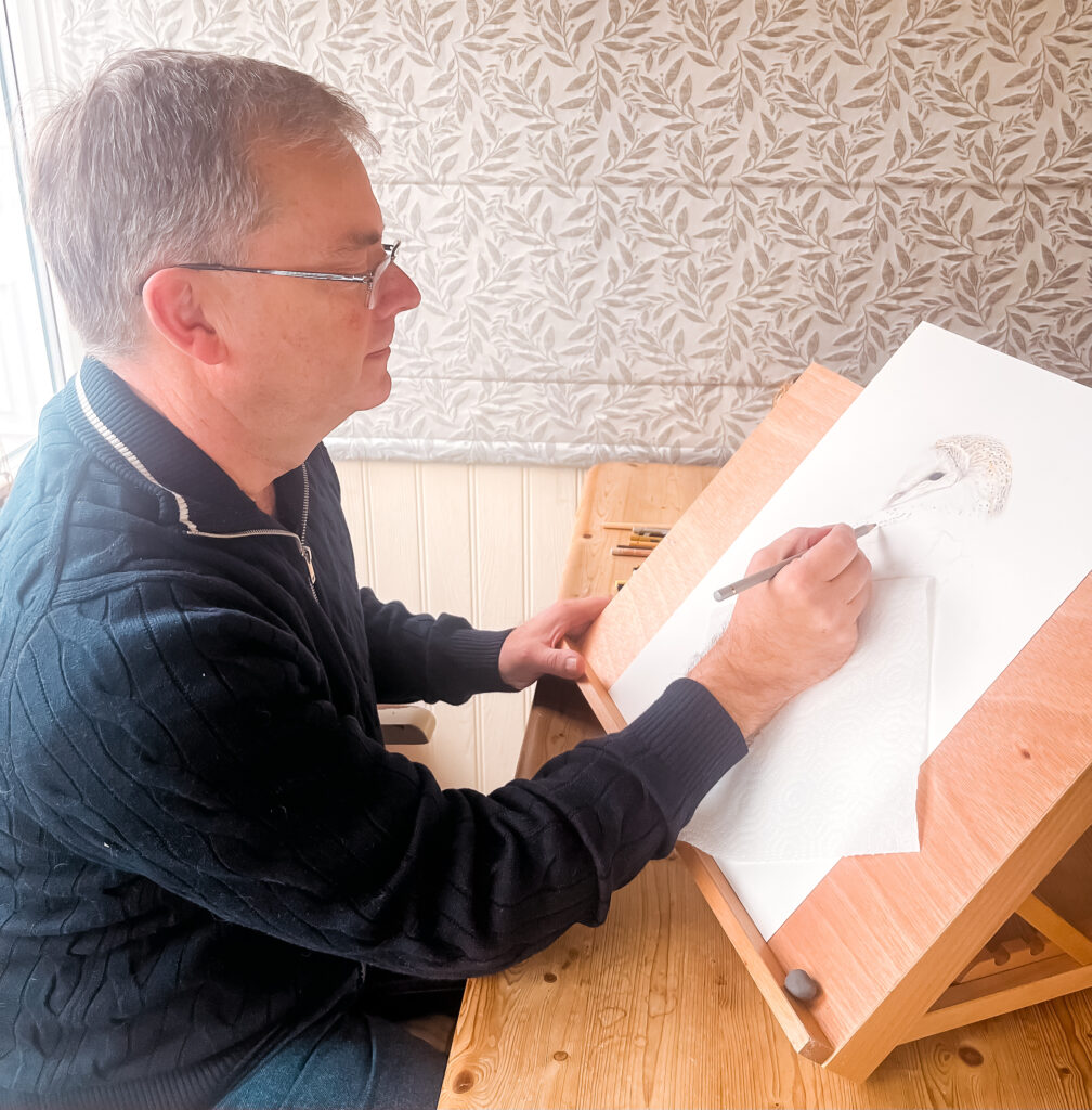Lee, a white man with short grey hair, is seen from the side drawing an owl with a pencil on a drawing board